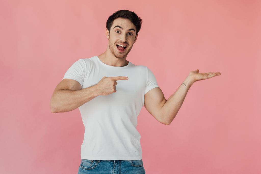 Aufgeregter muskulöser Mann im weißen T-Shirt, der mit der Hand zeigt und mit dem Finger isoliert auf rosa zeigt - Foto, Bild