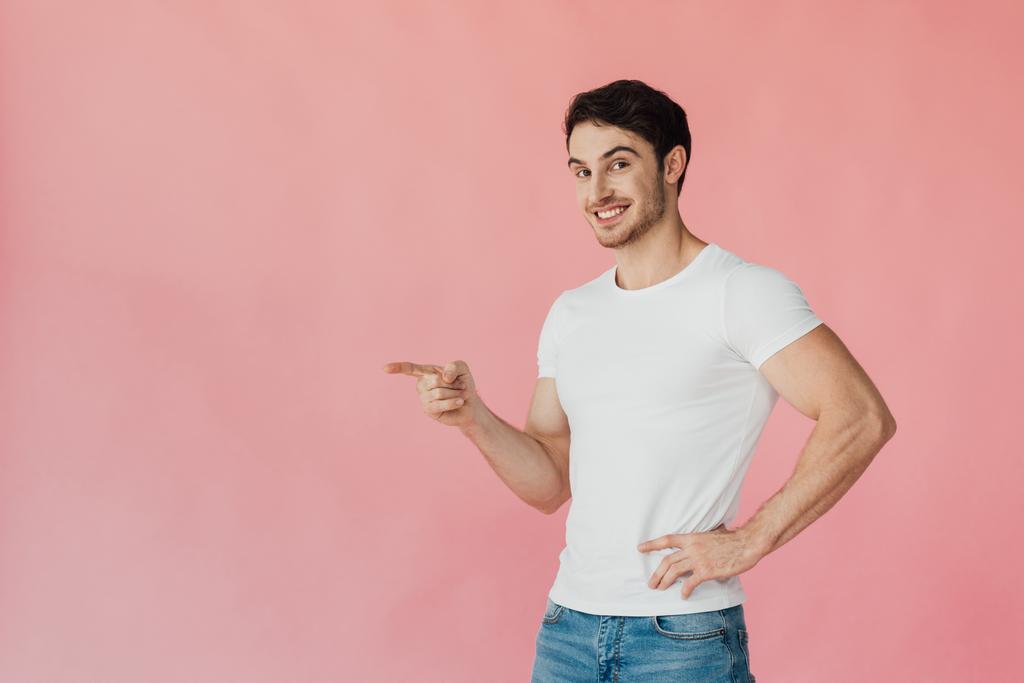 улыбающийся мускулистый мужчина в белой футболке, стоящий с рукой на бедре и указывающий пальцем, изолированным на розовый
 - Фото, изображение