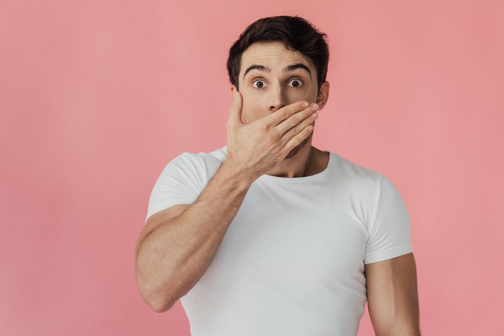 front view of shocked muscular man in white t-shirt covering mouth with hand isolated on pink - Photo, Image