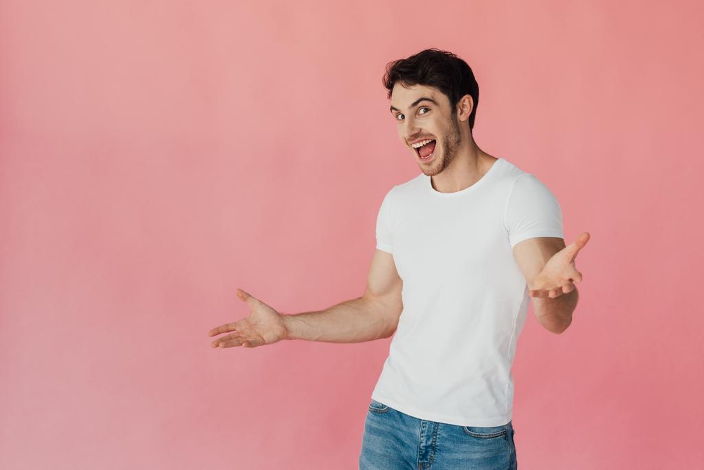 ενθουσιασμένος μυώδης άνθρωπος με λευκό μπλουζάκι που κουνώντας τα χέρια και κοιτάζοντας την κάμερα απομονώθηκε σε ροζ - Φωτογραφία, εικόνα