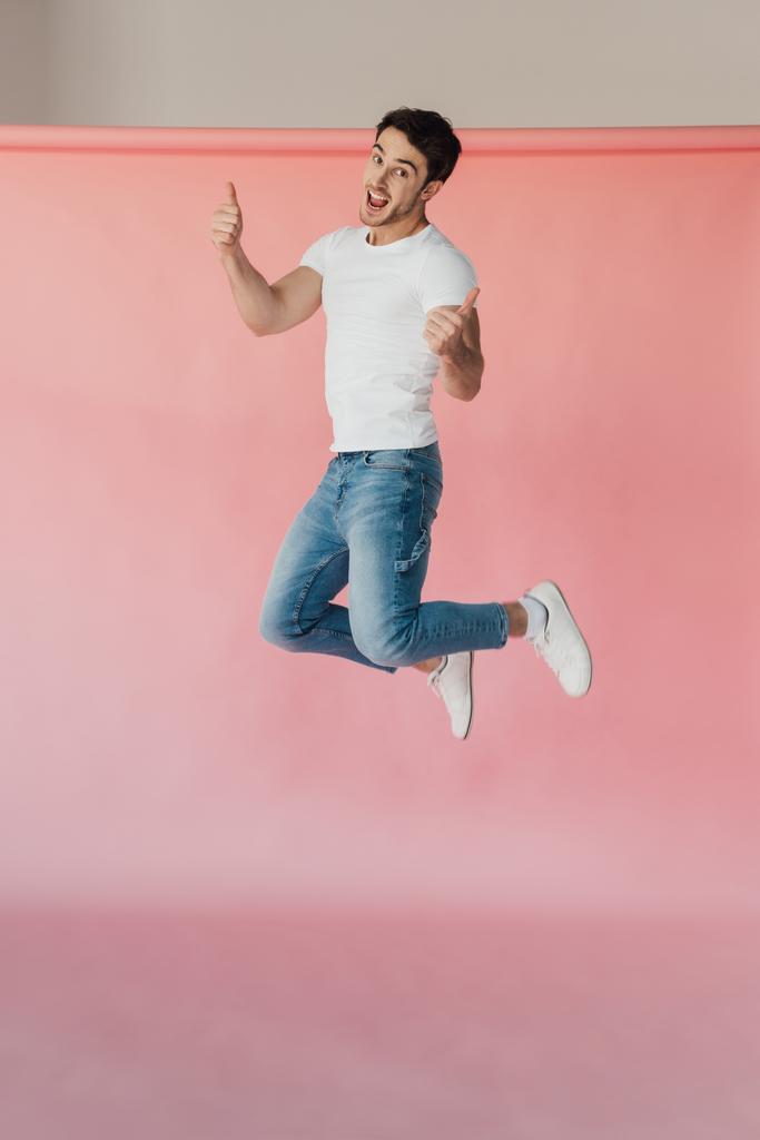 glimlachend gespierde man in wit t-shirt en jeans springen en tonen duimen omhoog op roze - Foto, afbeelding