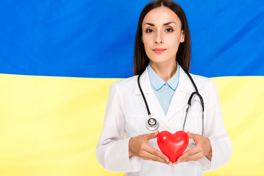 doctor in white coat with stethoscope holding red heart on Ukrainian flag background - Photo, Image