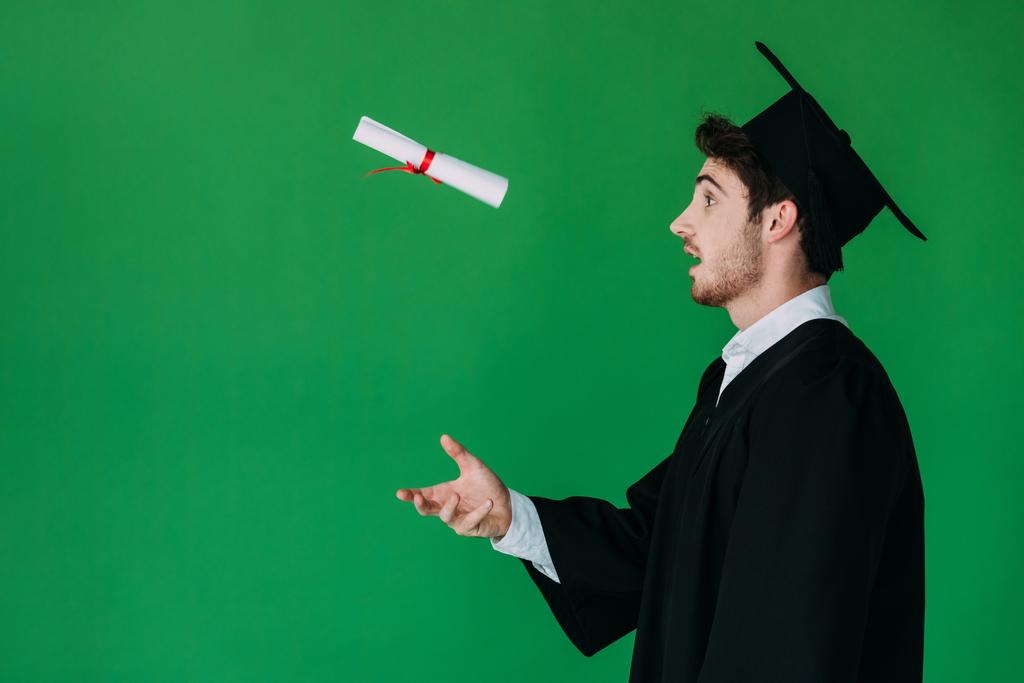 vista lateral del estudiante sorprendido en gorra académica vomitando diploma con cinta roja aislada en verde
 - Foto, Imagen