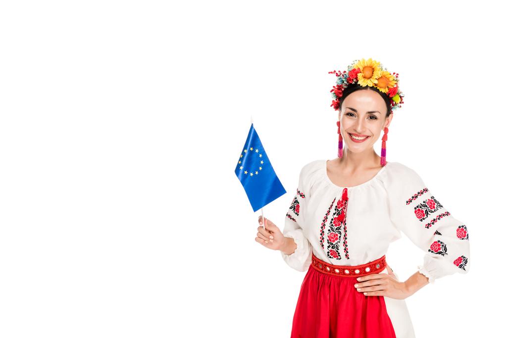 ευτυχής μελαχρινή νεαρή γυναίκα σε εθνικό ουκρανικό κοστούμι κρατώντας Ευρωπαϊκή σημαία απομονωμένη σε λευκό - Φωτογραφία, εικόνα