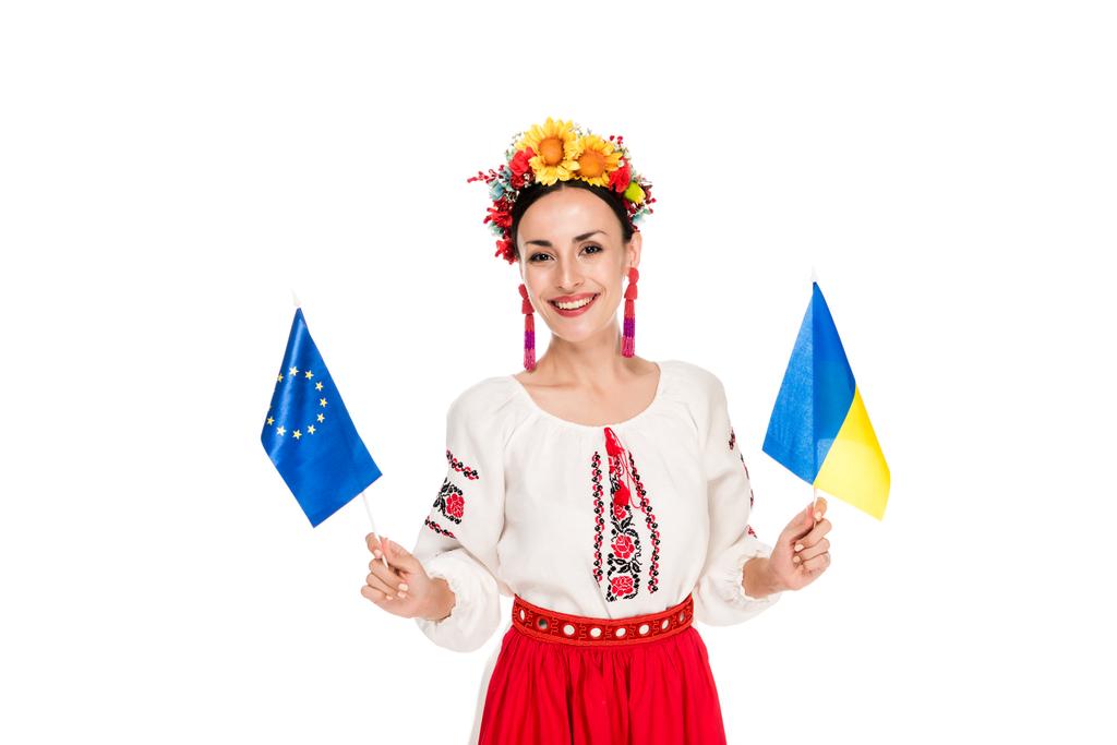happy brunette young woman in national Ukrainian costume holding European and Ukrainian flags isolated on white - Photo, Image