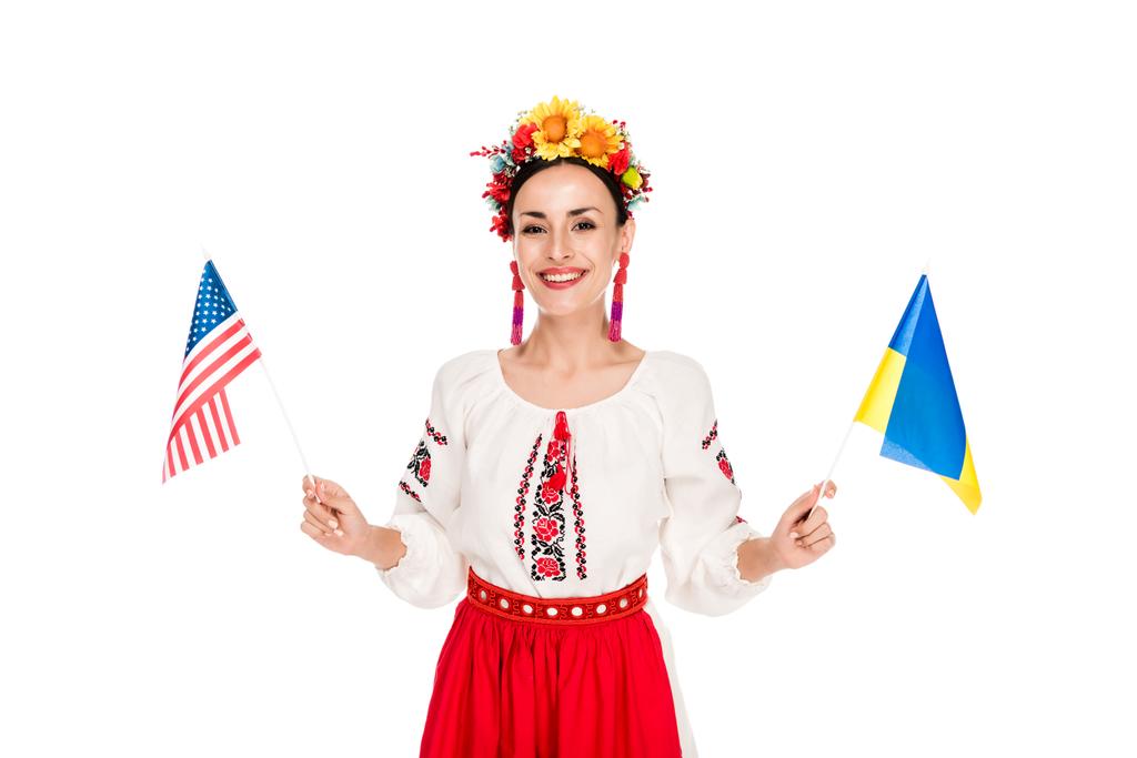 glimlachend brunette jonge vrouw in nationale Oekraïense kostuum houden van Amerikaanse en Oekraïense vlaggen geïsoleerd op wit - Foto, afbeelding