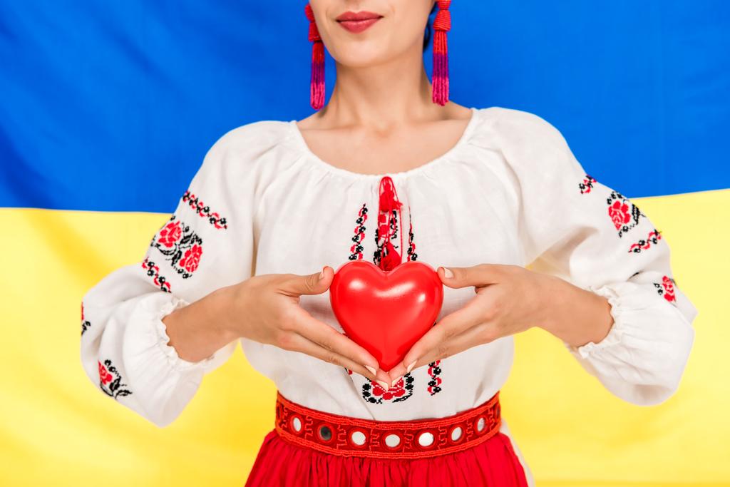arka planda Ukrayna bayrağı ile kırmızı kalp tutan ulusal Ukrayna kostümgenç kadının kısmi görünümü - Fotoğraf, Görsel