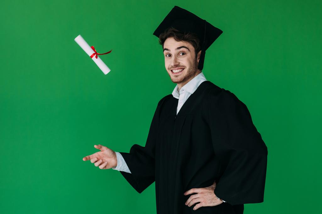 excited student in academic cap throwing up diploma with red ribbon isolated on green - Photo, Image