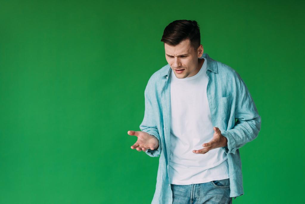 Раздражённый молодой человек в рубашке жестикулирует изолированным на зеленом
 - Фото, изображение