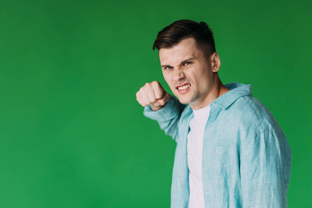 angry young man in shirt holding fist up and looking at camera isolated on green - Photo, Image