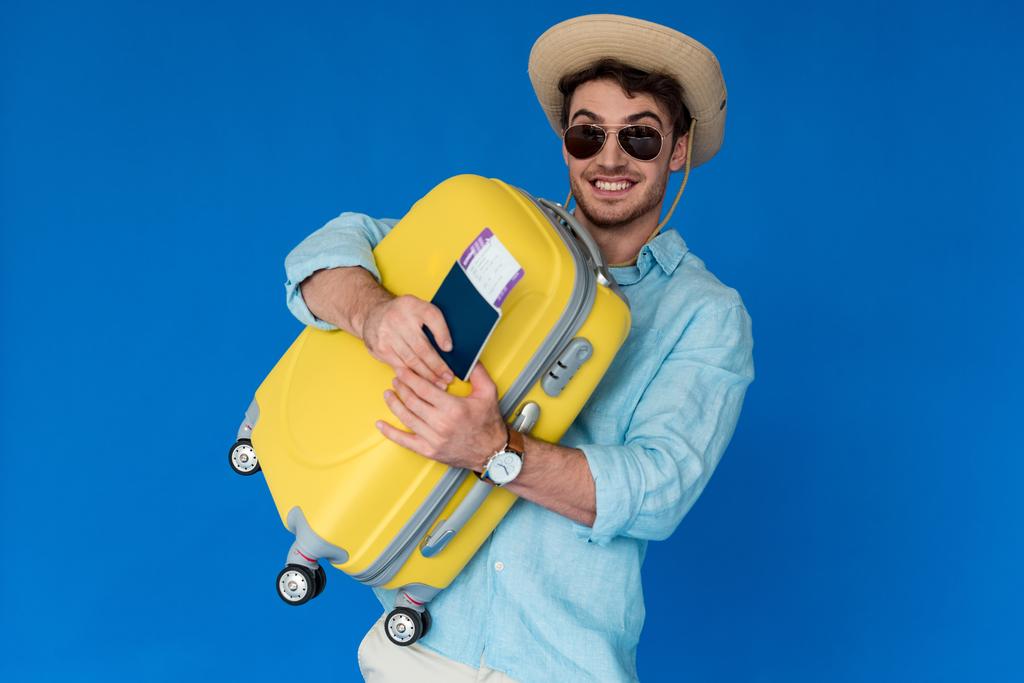 podekscytowany podróżnik w Safari kapelusz i okulary przeciwsłoneczne trzymając żółty walizkę i paszport z biletem lotniczym na niebiesko - Zdjęcie, obraz