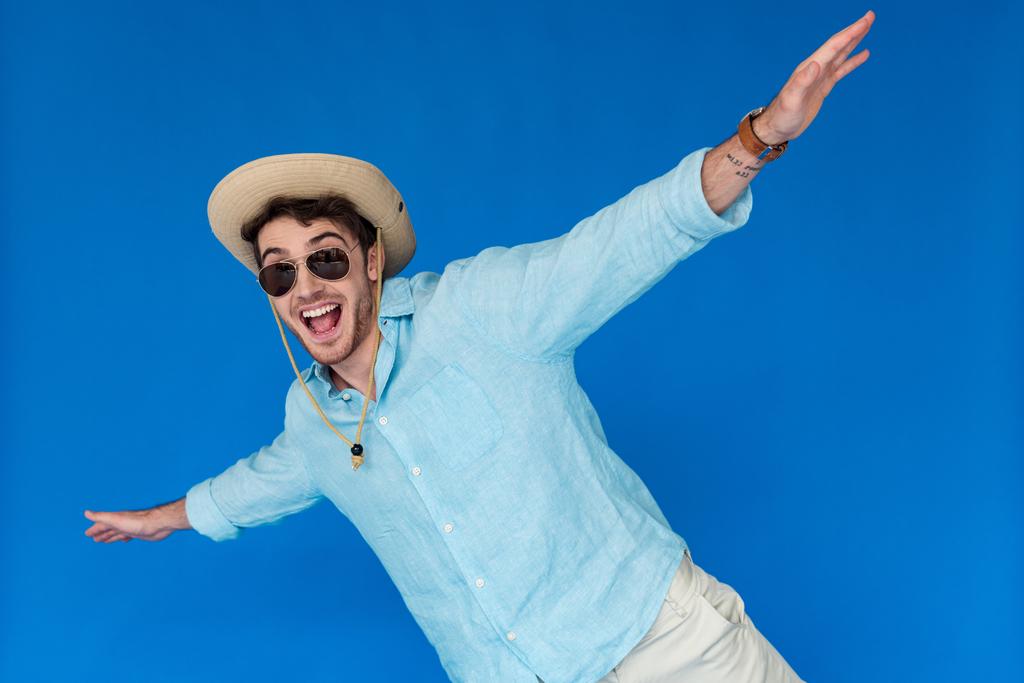 Aufgeregter Tourist mit Safarimütze und Sonnenbrille, lachend und winkend mit den Händen auf blauem Grund - Foto, Bild