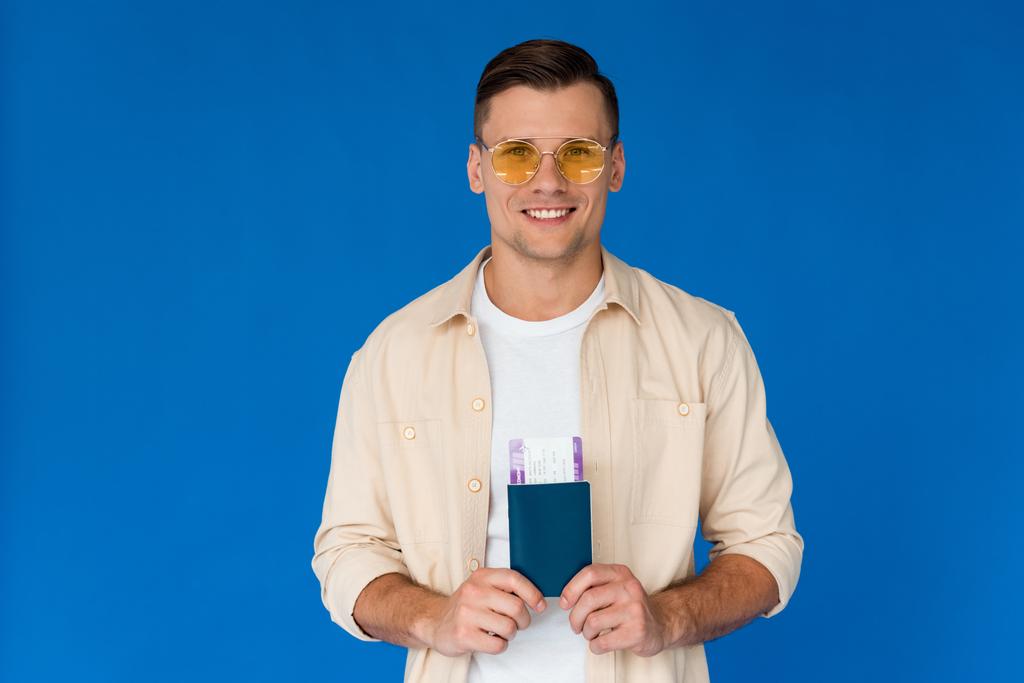 青で隔離された航空券でパスポートを持つサングラスをかけた笑顔の旅行者の正面図 - 写真・画像