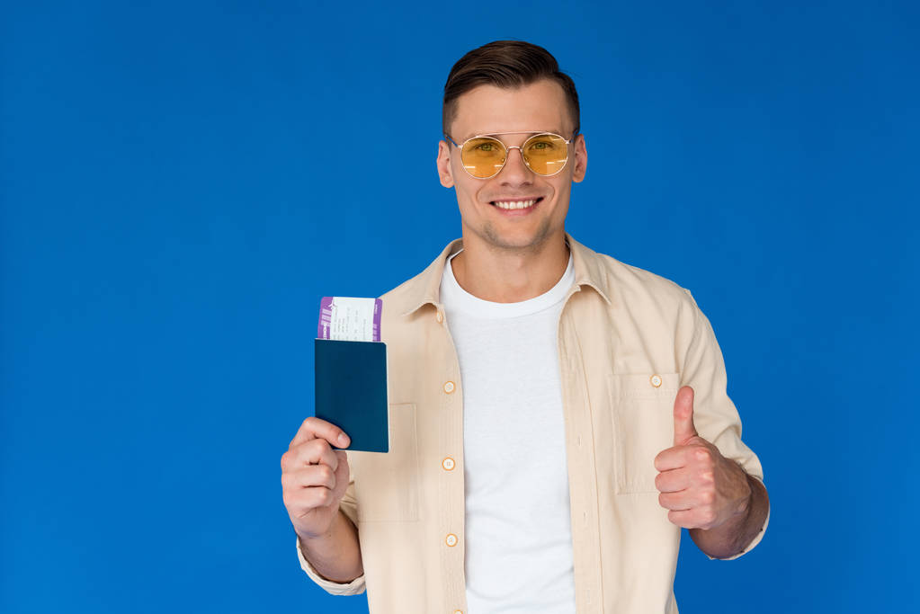 front view of smiling tourist in sunglasses holding passport and showing thumb up with air ticket isolated on blue - Photo, Image