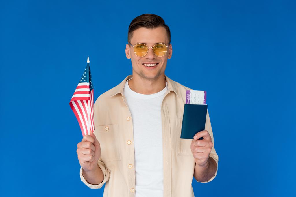 Vooraanzicht van lachende toerist in zonnebril houden paspoort met vliegticket en Amerikaanse vlag geïsoleerd op blauw - Foto, afbeelding
