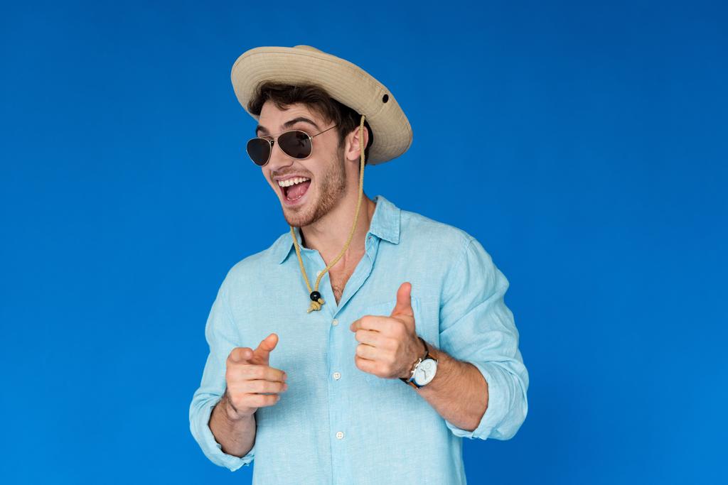 usměvavý cestovatel v Safari klobouku ukazující palec nahoru a ukazování prstem na kameru izolovaně na modré - Fotografie, Obrázek