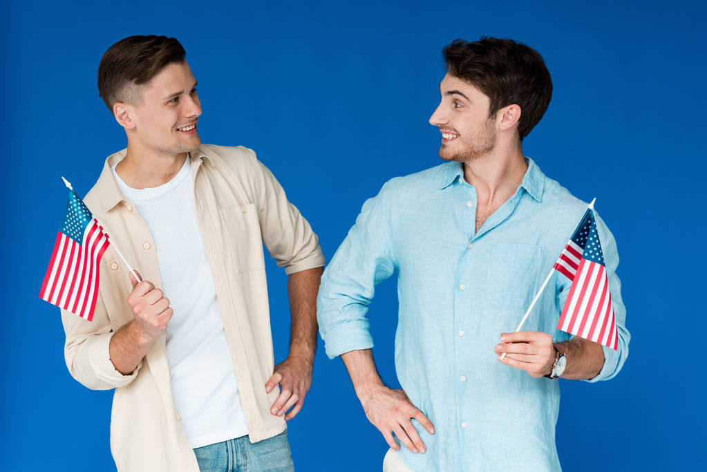 two smiling friends holding american flags and looking at each other isolated on blue - Photo, Image