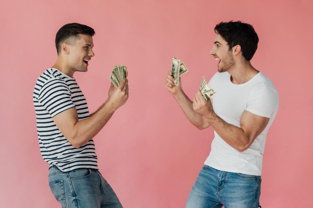 dva nadšené přátele s dolarovou bankovkami, kteří se na sebe dívali izolovaně na růžovém - Fotografie, Obrázek