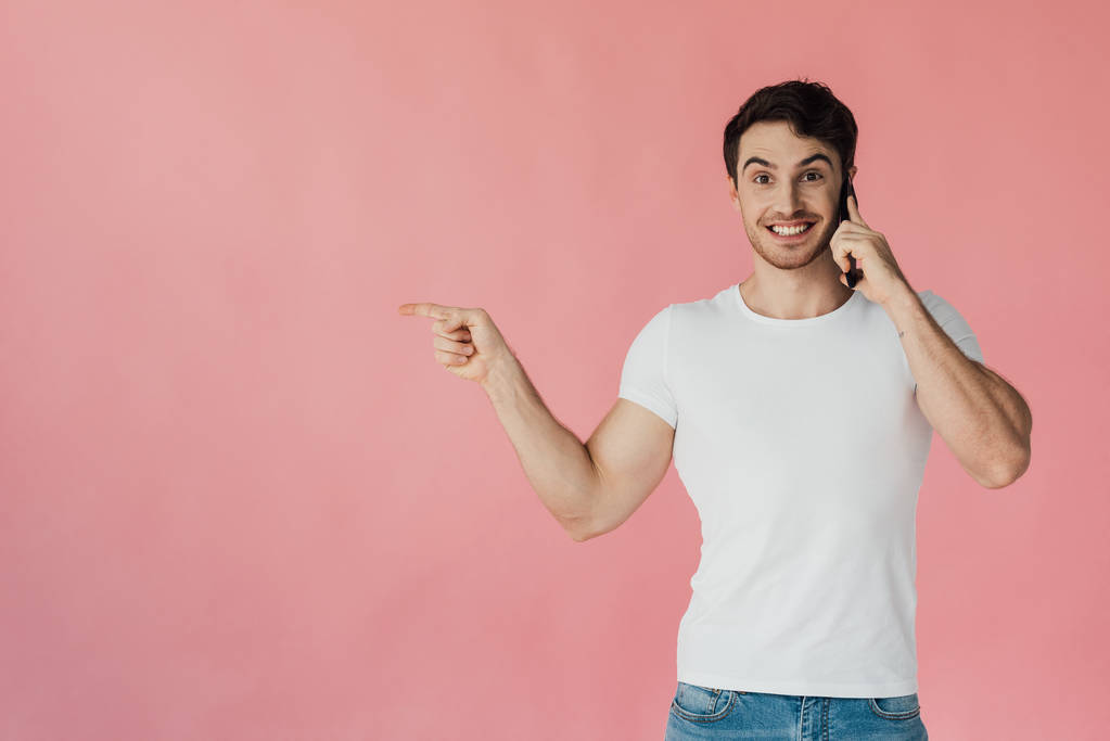 Vorderansicht eines lächelnden muskulösen Mannes in weißem T-Shirt, der auf dem Smartphone spricht und mit dem Finger isoliert auf rosa zeigt - Foto, Bild