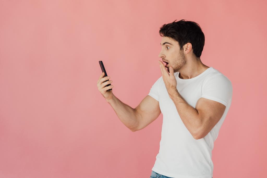 bang gespierde man in wit t-shirt houden smartphone geïsoleerd op roze - Foto, afbeelding