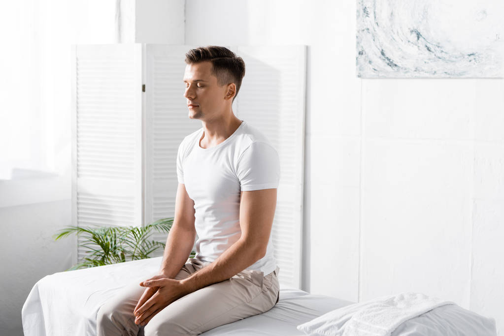 hombre en camiseta blanca sentado en la mesa de masaje con los ojos cerrados en la clínica
 - Foto, imagen