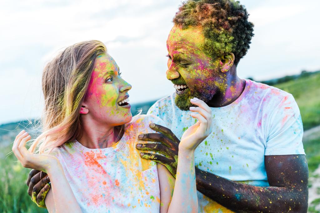 happy african american man touching shoulders of surprised young woman with holi paints on face  - Photo, Image