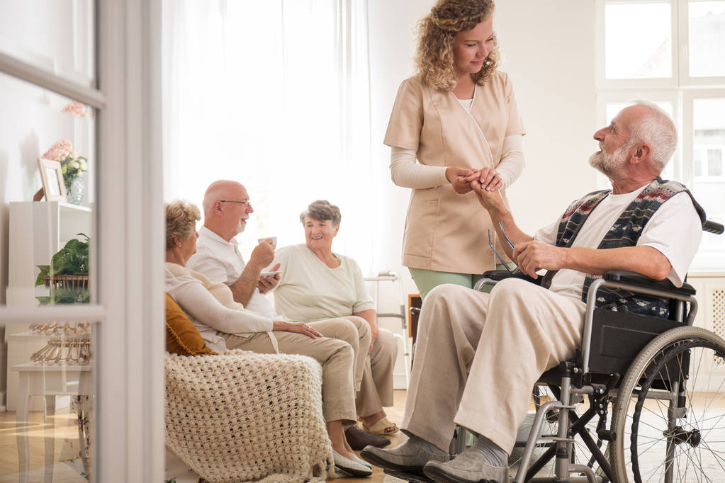 Hombre mayor en silla de ruedas con una enfermera servicial sosteniendo su mano y amigos sentados en el sofá bebiendo té
 - Foto, imagen