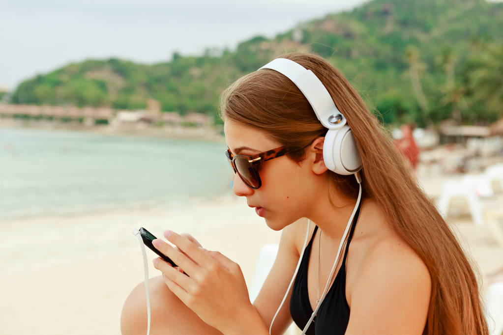 Portret młodej dziewczyny Piękna Nastolatka siedzi białym piaskiem plaży słuchania muzyki za pomocą słuchawek i smartfona przeciw na tle zieleni. Młodzieńczy styl życia na zewnątrz. - Zdjęcie, obraz