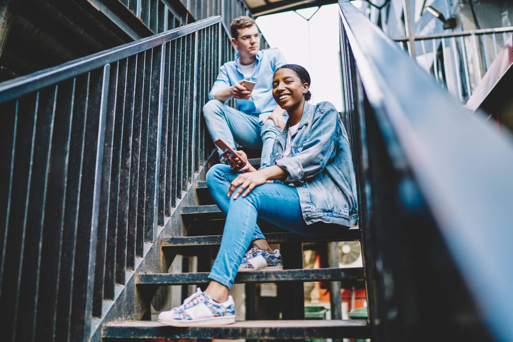 明るいアフリカ系アメリカ人の若い女性の肖像は、階段に白人の友人と座っているスマートフォンのアプリ上のプロファイルを更新しながら、カメラで笑顔. - 写真・画像