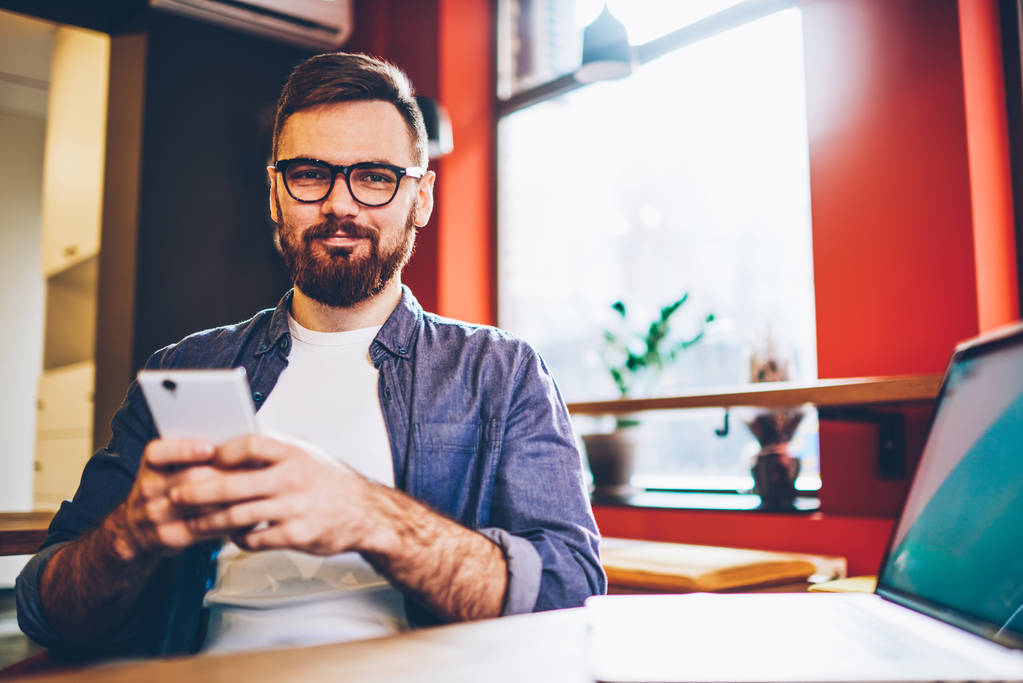 Portrait of handsome bearded male blogger looking at camera while spending leisure time in cafe interior with good internet connection, young hipster guy in eyeglasses holding smartphone gadget - Photo, Image