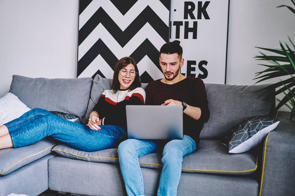 Caucasian man booking tickets on website via laptop sitting on comfortable couch with girlfriend using home internet connection, couple in love shopping together on computer while resting on sofa - Photo, Image