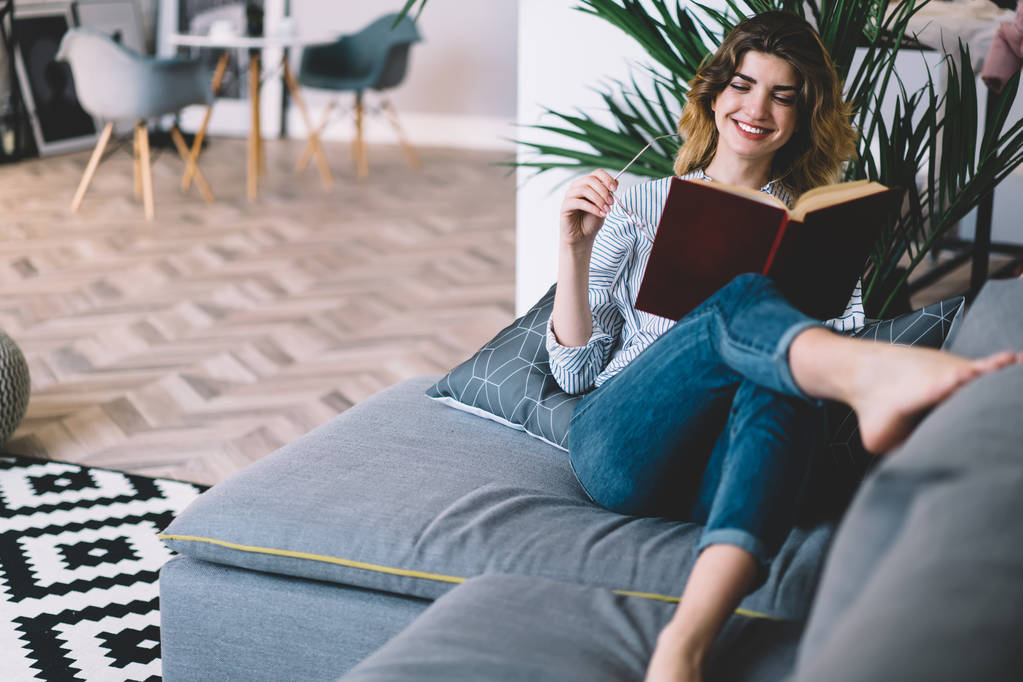Positive young woman happy with recreation time at home enjoying interesting book with funny plot.Cheerful hipster girl lying on comfortable couch and reading textbook in modern apartment - Valokuva, kuva