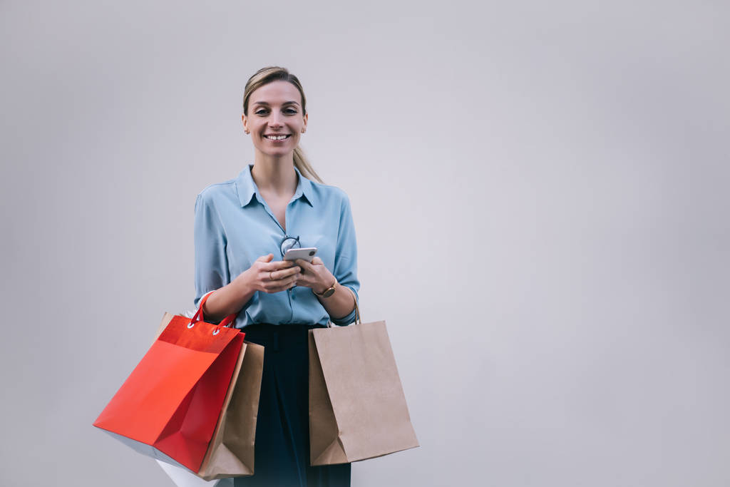 Портрет позитивной женщины в стильном наряде, держащей в руках мобильный телефон и бумажные пакеты и улыбающейся перед камерой, веселой миллениалки, стоящей на рекламном фоне - Фото, изображение