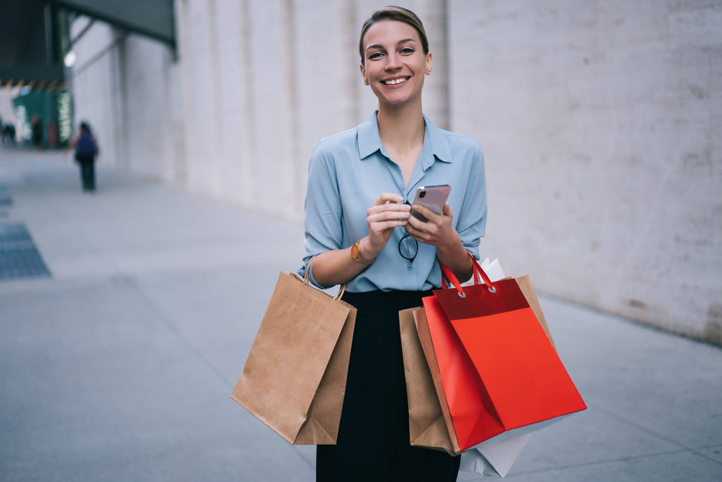Poloviční portrét vzrušeného ženského kupce stojící na reklamní ploše s papírovými obaly a usmívající se na kameru, šťastná hipsterová dívka v módním oblečení držící moderní mobilní přístroj venku - Fotografie, Obrázek
