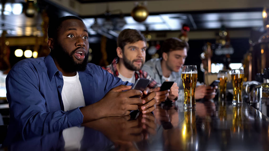 Homem afro-americano verificando apostas no smartphone, assistindo jogo no bar, app
 - Foto, Imagem