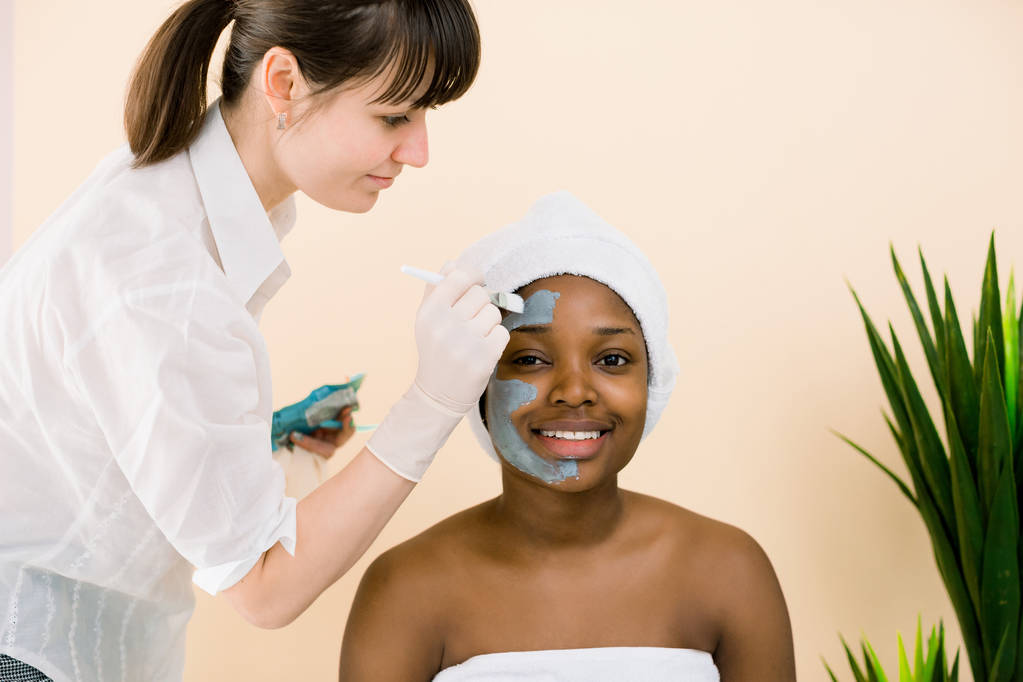 スパサロンでアフリカ系アメリカ人女性の顔にマスクを塗る美容師。スパでフェイシャルマスクを手に入れる若いアフリカの女性 - 写真・画像