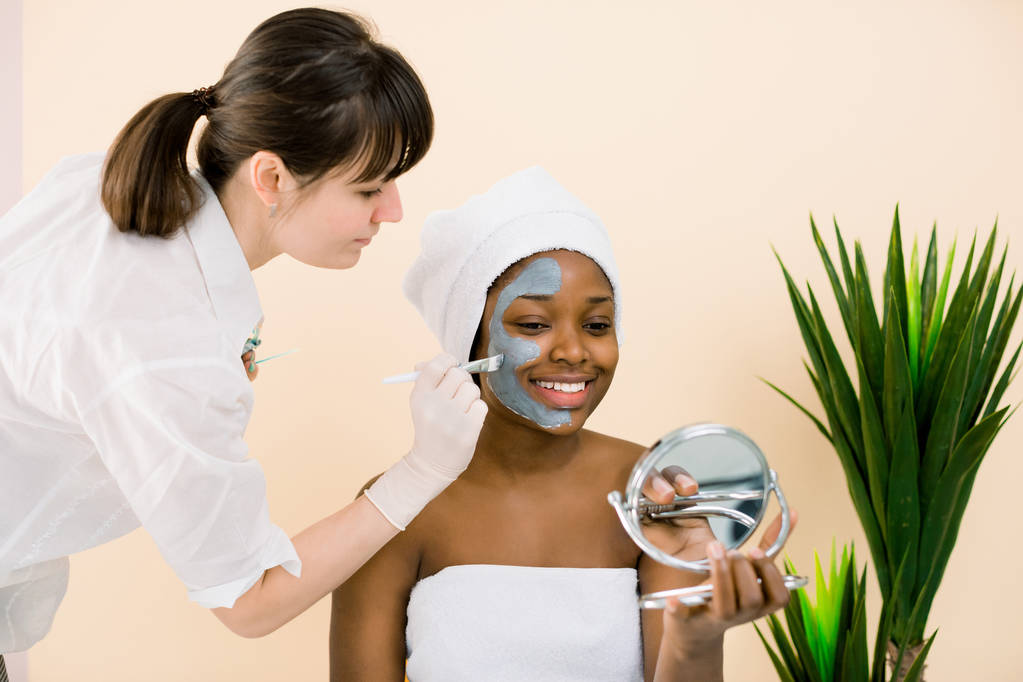 顔のルーチンの間に白いタオルでかなりイプンアフリカの女性は、マスクを適用し、鏡で見て。スパサロンでアフリカ系アメリカ人女性の顔にマスクを塗る美容師 - 写真・画像