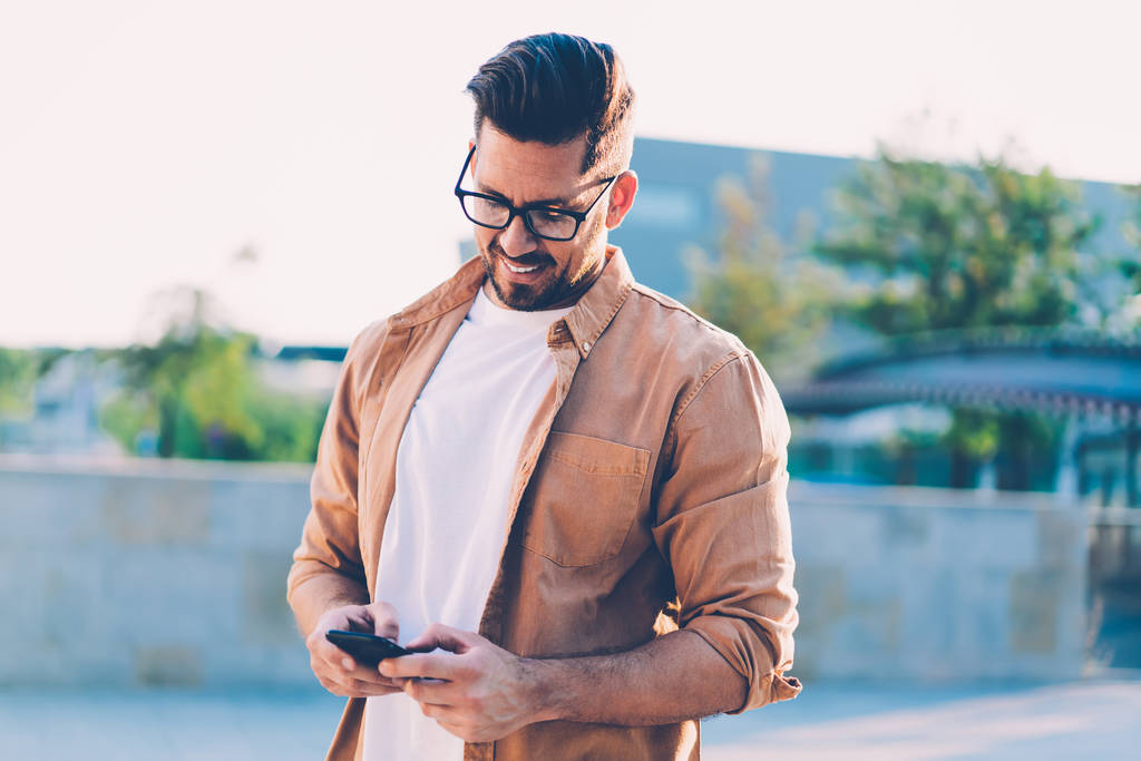 Erfolgreicher Mann mit optischer Brille überprüft Benachrichtigung für mobile Anwendung in der Freizeit auf städtischem Gelände, glücklicher Mann in lässigem Hemd sendet SMS-Nachricht während der Online-Kommunikation - Foto, Bild