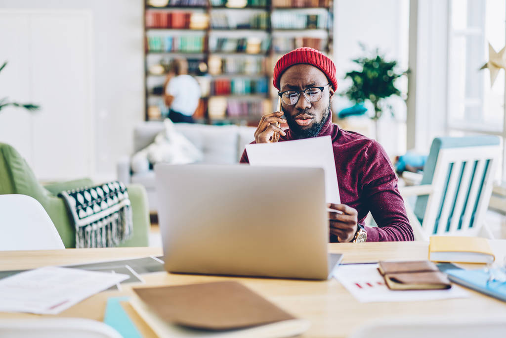 Serious african american hipster guy reading document and having mobile phone conversation doing remote job at home,male freelancer talking on cellular about online business holding paper with plan - Photo, Image