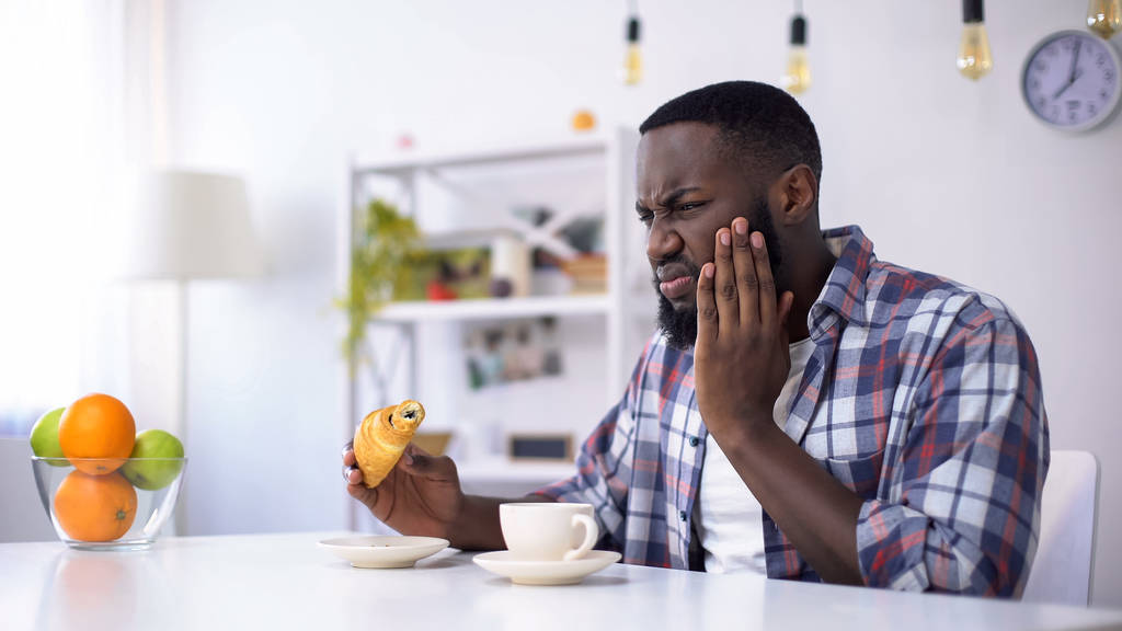 schwarzer Mann, der Zahnschmerzen durch süßes Essen verspürt, Croissant mit Kaffee isst - Foto, Bild