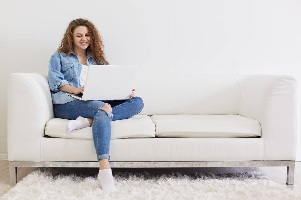 Fotó a bájos szép elégedett hölgy göndör hajú ül a fehér kényelmes kanapé, a laptop segítségével az online munka, vezeték nélküli internet, hogy a világos belső szobában, úgy néz ki, boldog és boldog. - Fotó, kép