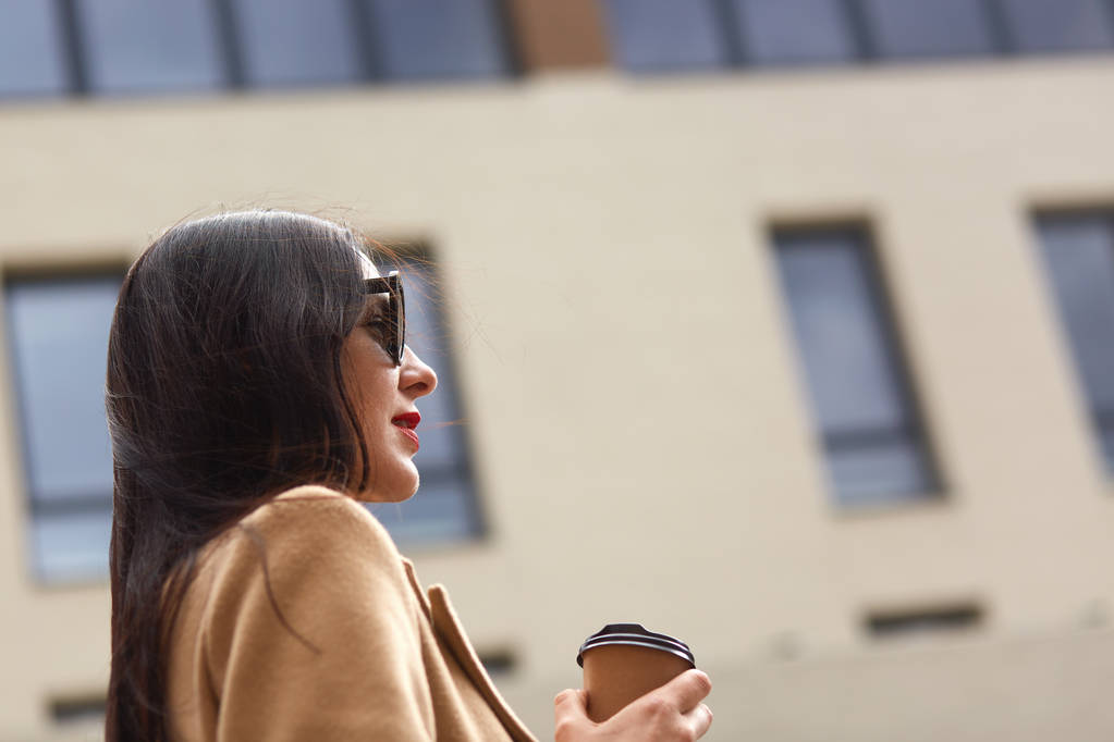 Retrato lateral de joven morena turista visitando la ciudad y sosteniendo taza de café para llevar de papel mientras mira a la distancia a través de gafas de sol con estilo negro, teniendo recreación en otra ciudad
. - Foto, imagen