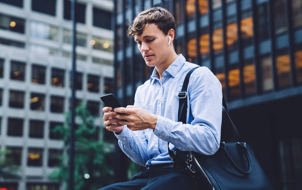 Muž podnikatel ve sluchátkách s formálním kufříkem sedí na městském prostředí čeká obchodní partner ve finanční čtvrti pomocí 4g bezdrátové připojení k internetu na mobilní telefon gadget pro psaní sms - Fotografie, Obrázek