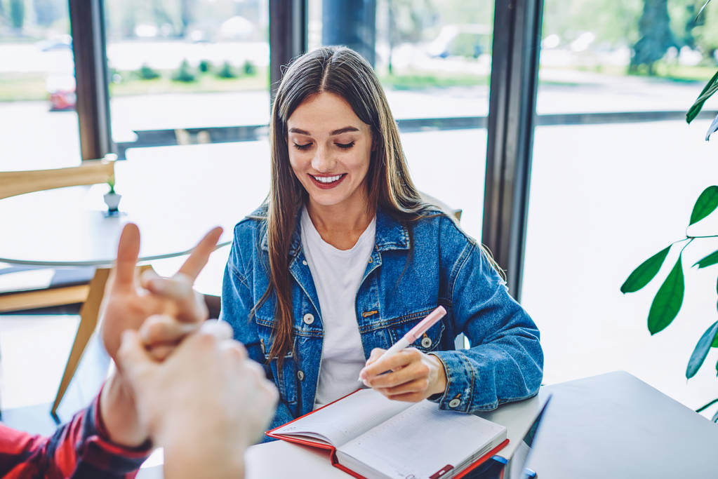 Happy woman making notes of organization plan in textbook during leisure time with friend sitting near, positive hipster girl preparing to university examination with notebook for education. - Foto, Imagen