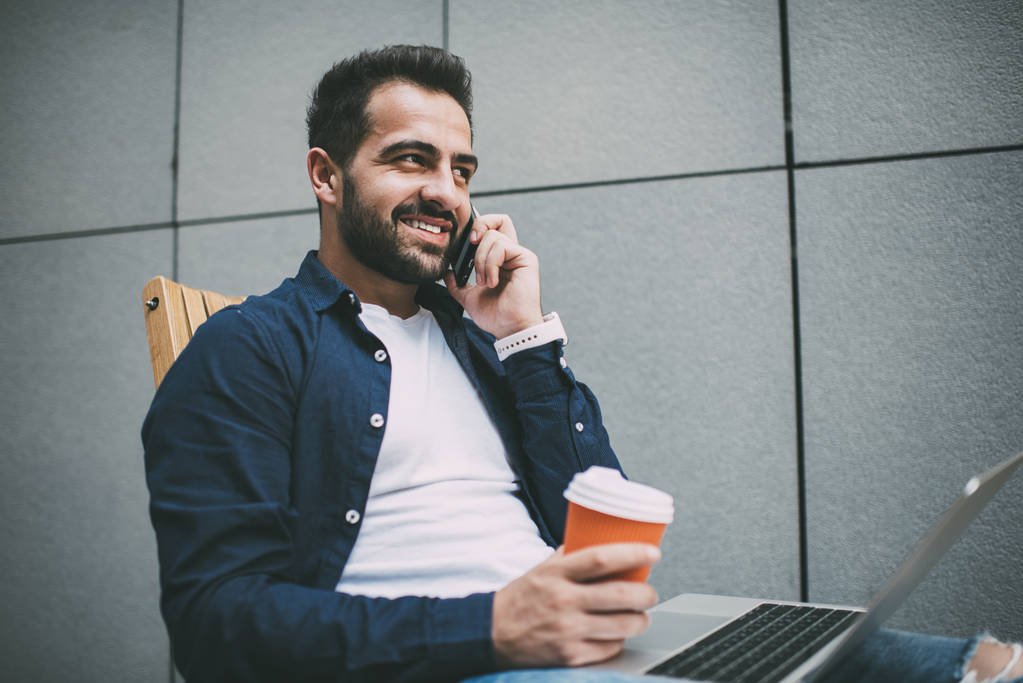 Sukces brodaty freelancer dzwoni do znajomego w celu komunikacji podczas przerwy na kawę z programowania online przez netbook, pozytywny hipster facet dzwoni przez telefon komórkowy za pomocą roamingu 4g na zewnątrz - Zdjęcie, obraz