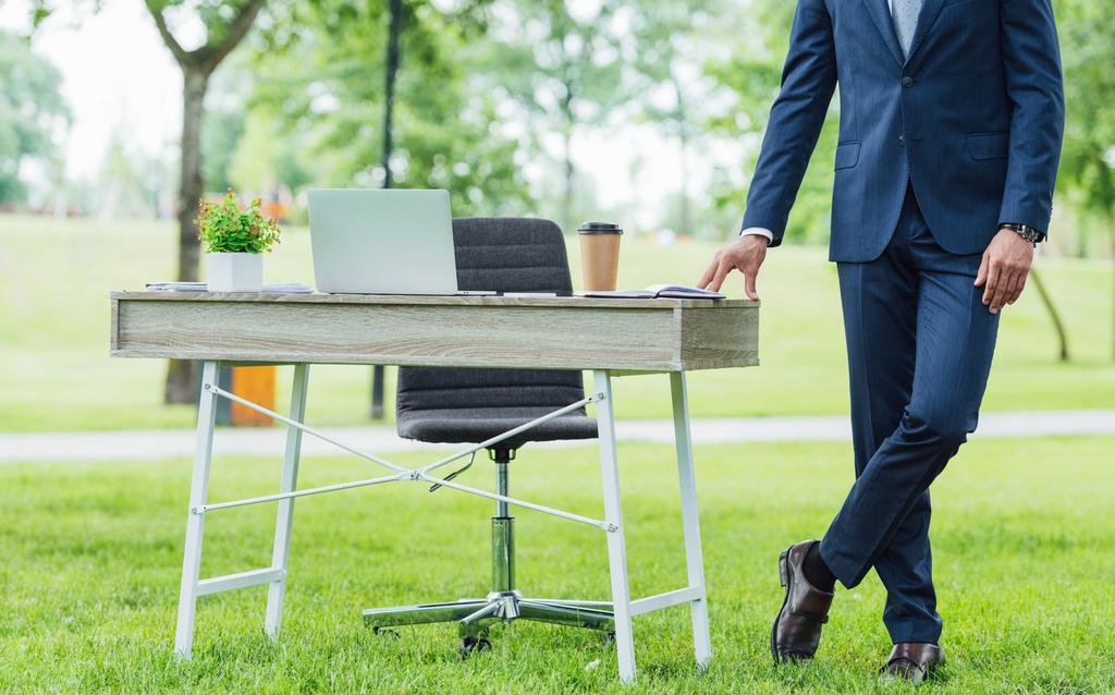 обрізаний вид молодого бізнесмена, що стоїть з схрещеними ногами біля столу в парку
 - Фото, зображення