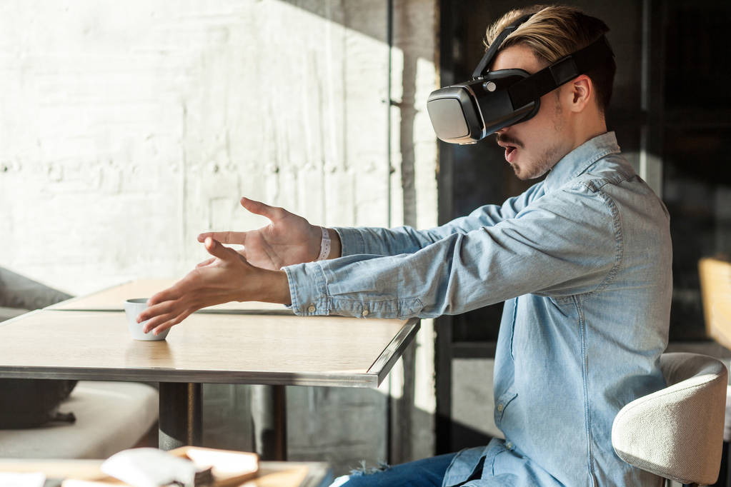 Zijaanzicht van verrast bebaarde man in blauw denim shirt met VR-headset en het bekijken van video op Simulator terwijl zittend met verhoogde armen en opende mond in café, Entertainment Lifestyle. - Foto, afbeelding