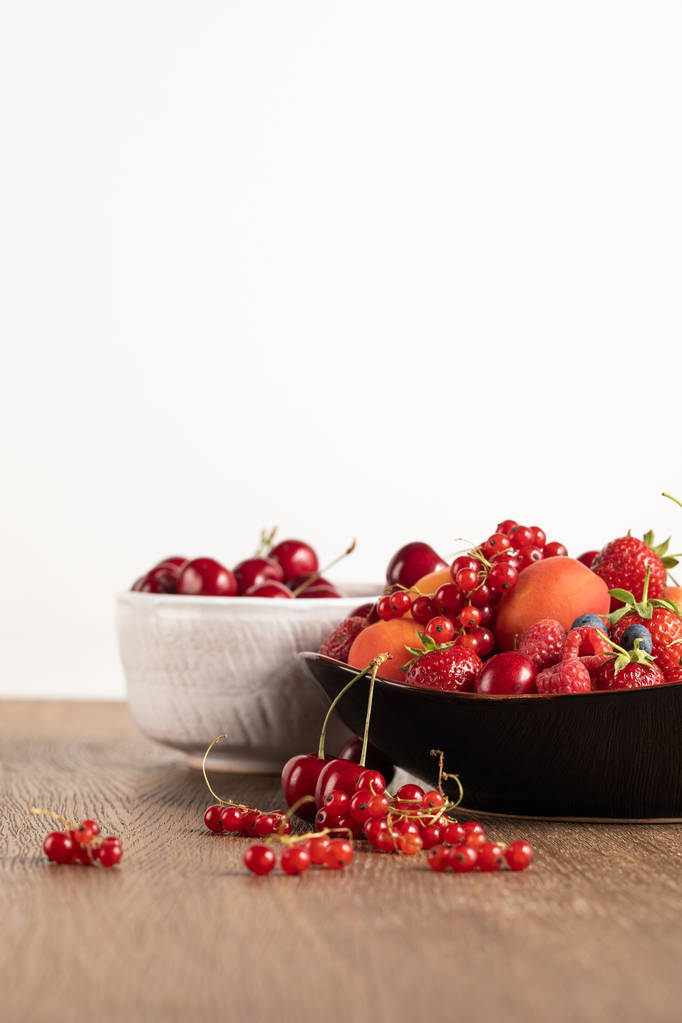 selektiver Fokus von roten Kirschen in weißer Schüssel und gemischten Beeren auf Teller auf Holztisch isoliert auf weißer - Foto, Bild