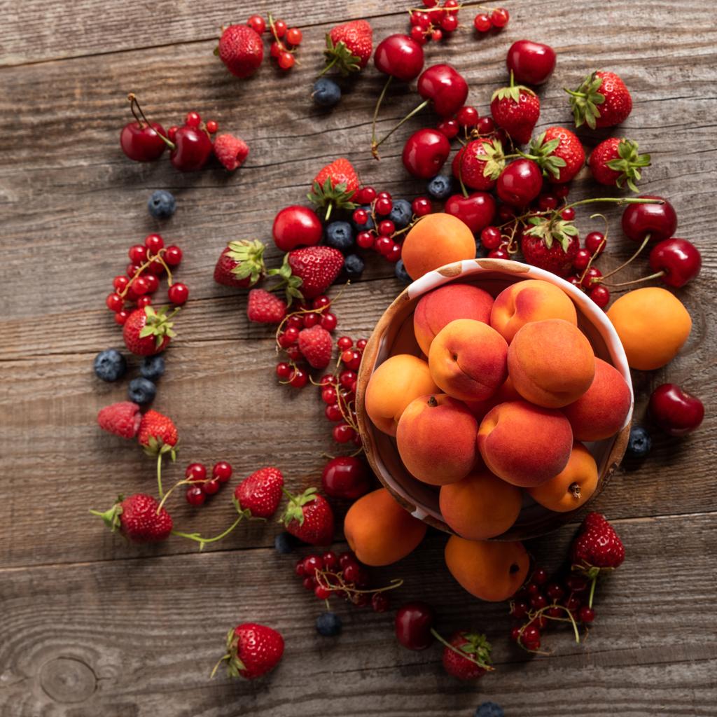 az érett, ízletes, szezonális bogyós gyümölcsök a tálból, fából készült asztal friss kajszibarack körül szétszórva  - Fotó, kép