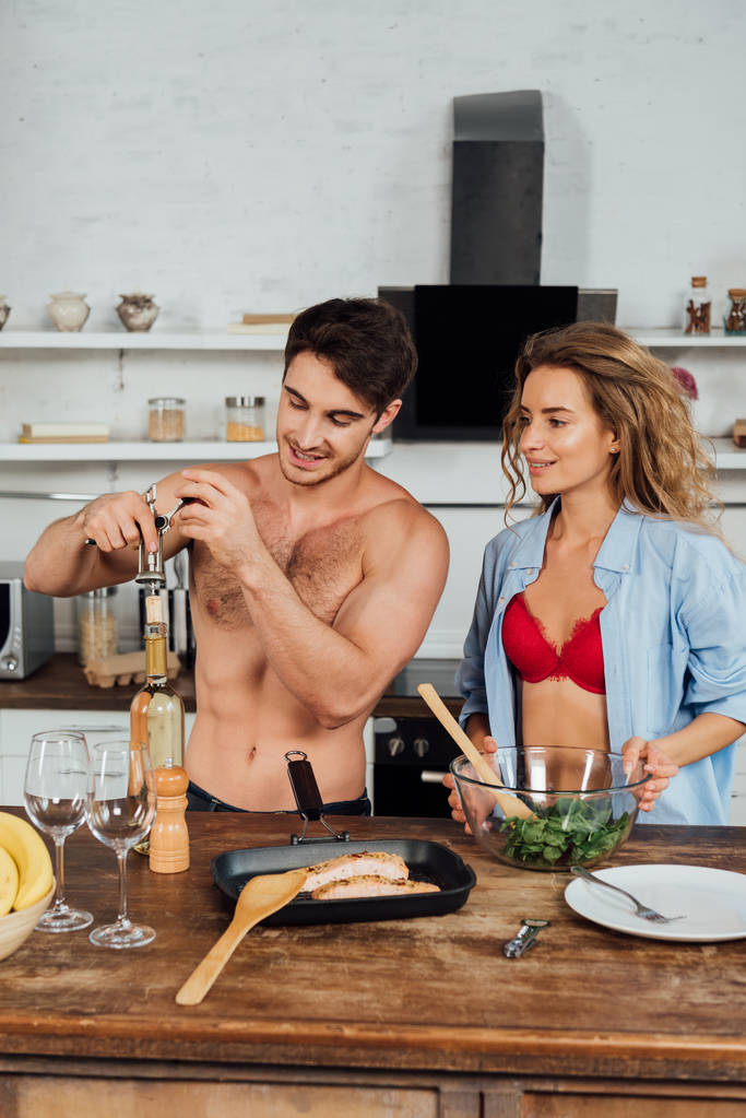 sexy Mädchen und hemdloser Mann öffnet Flasche Wein in der Küche - Foto, Bild
